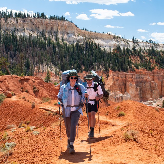 Two hikers along a trail surrounded by red rock slopes and towering rock spires