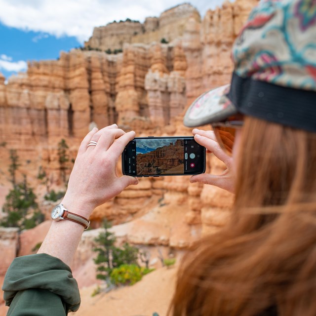 A woman holds a smartphone to take a photo of a redrock landscape