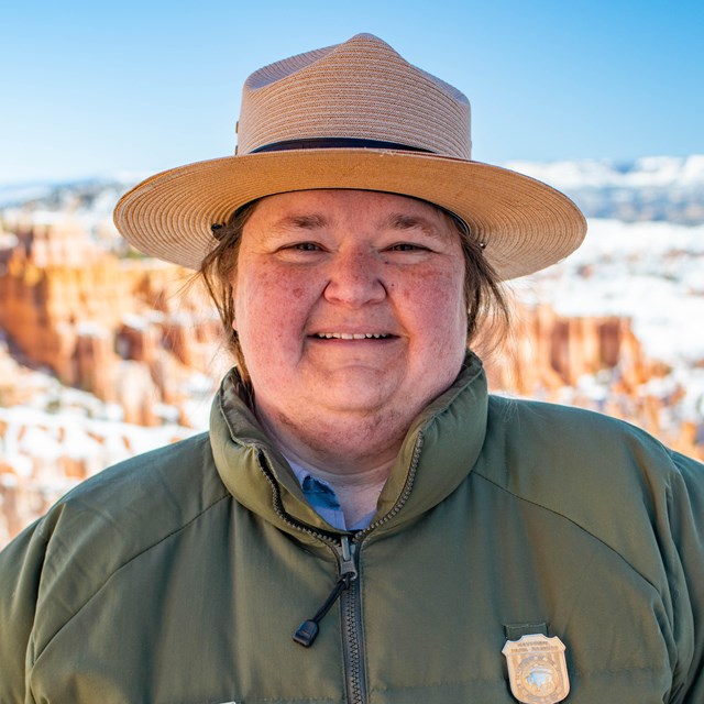 A woman in a National Park Service uniform and flat hat stands in front of red rocks and a blue sky