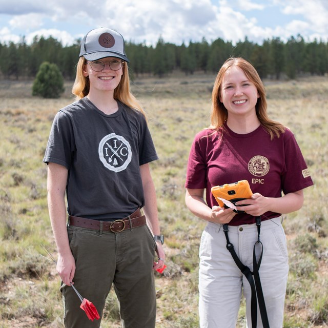 Two women stand in a field holding archaeology equipment.