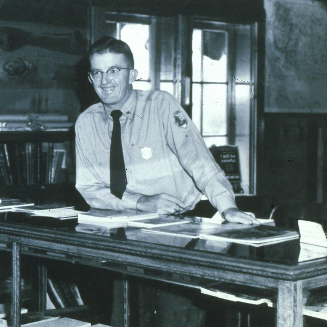 A black and white photo of a man in a park ranger uniform standing behind a desk