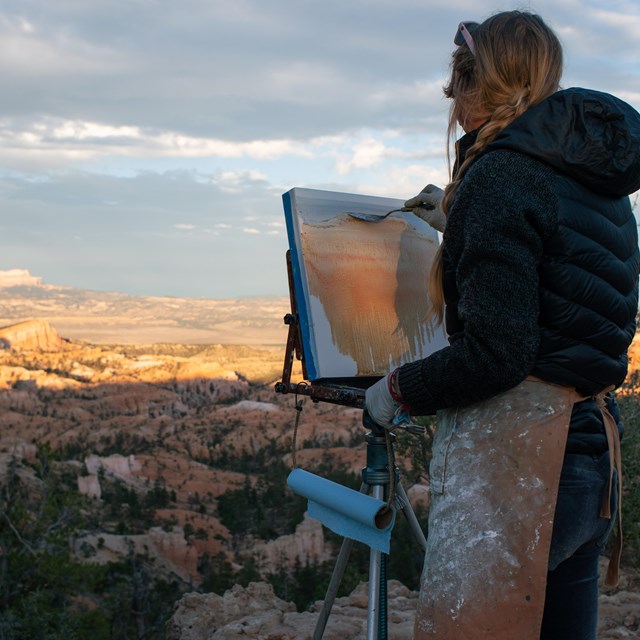 A woman stands on the rim of the canyon painting a canvas on an easel
