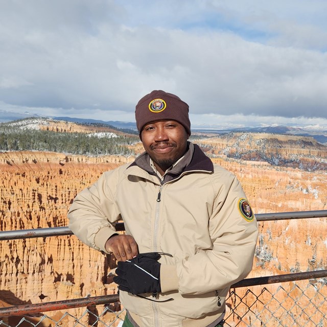 A man in winter clothing stands at the edge of a vast landscape of red rock spires.