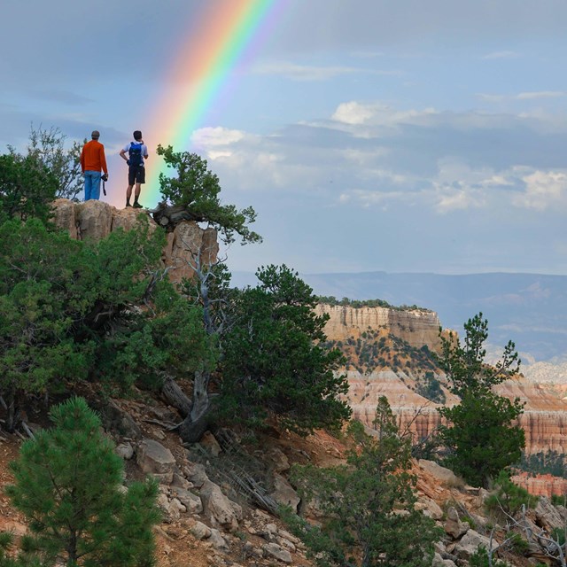 Two people stand along a trail viewing a rainbow