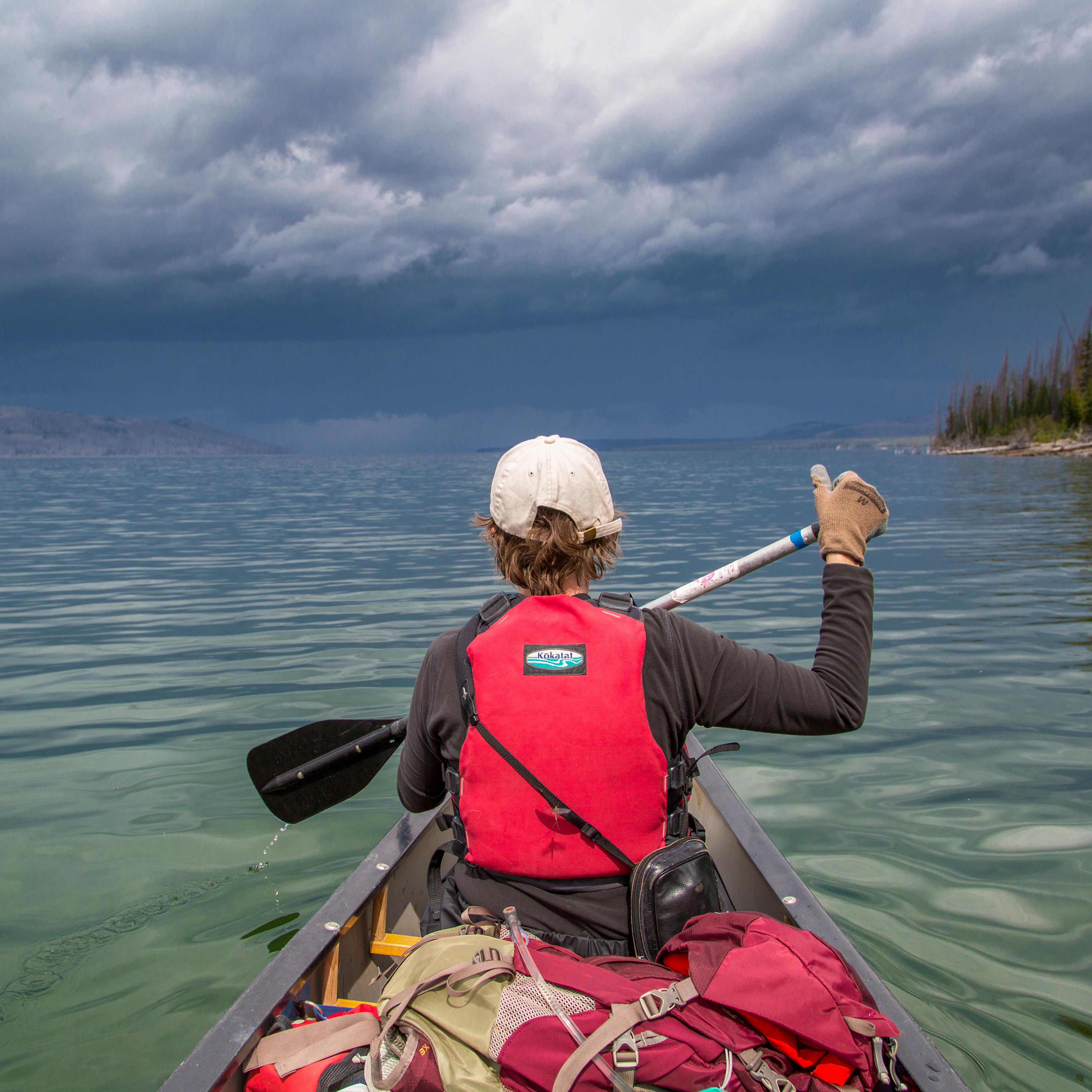 a boy in a red lifejacket paddles a canoe on a lake
