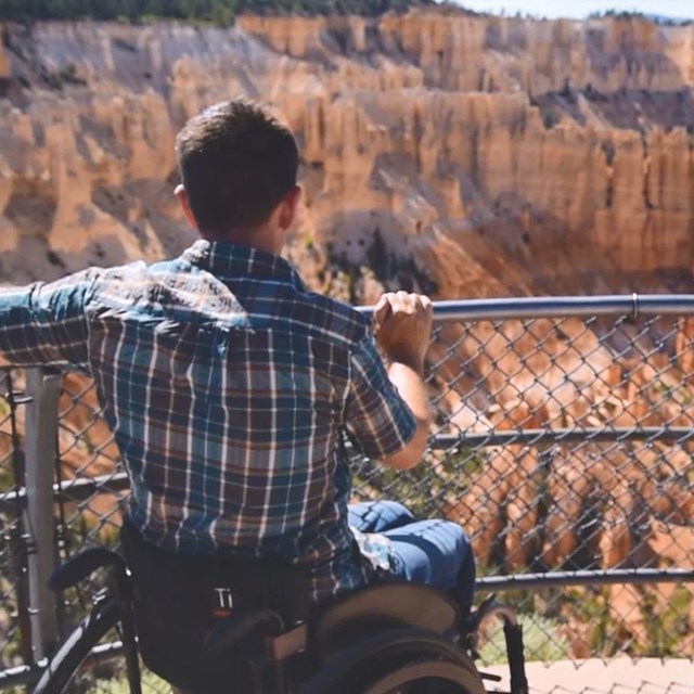 Man in wheelchair at railing overlooking vast landscape of red rock spires and cliffs.