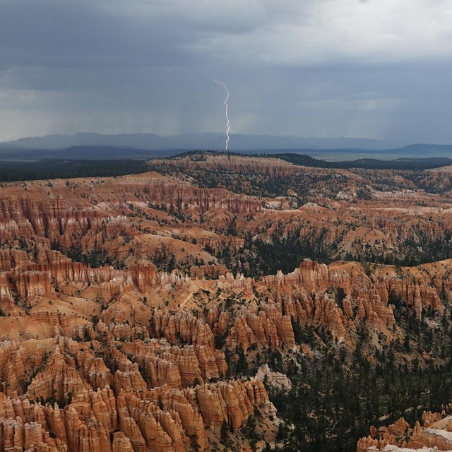A vast redrock landscape filled with rock spires and forest with dark skies and lightning above
