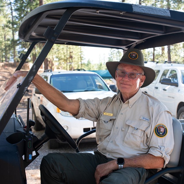 A man sits in a golf cart wearing a national park service volunteer hat and shirt.