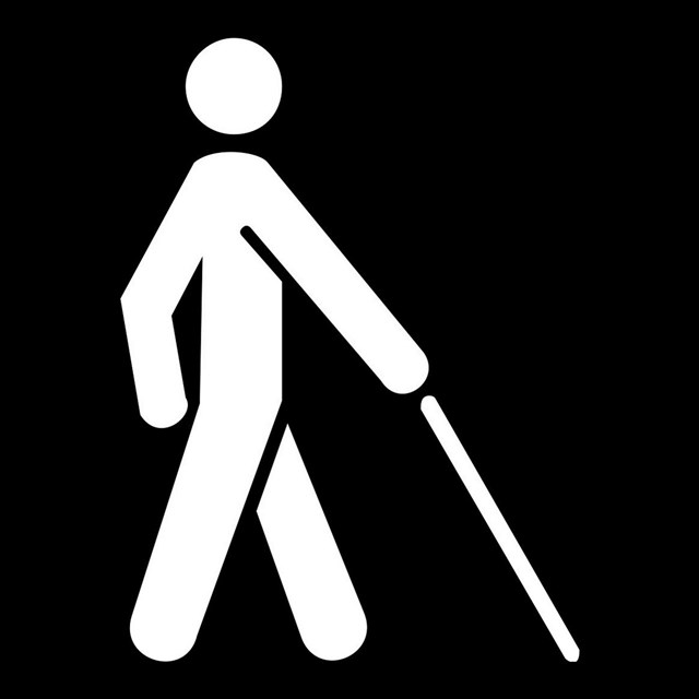 A black and white icon of a person with a seeing-eye cane.