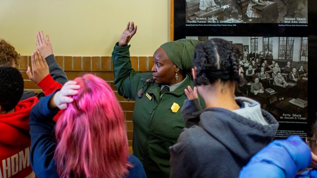 A park ranger raises her hand along with a group of children in front of a photo of discrimination.
