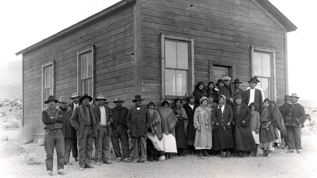 Several Native Americans standing in front of a small wooden building in the desert.