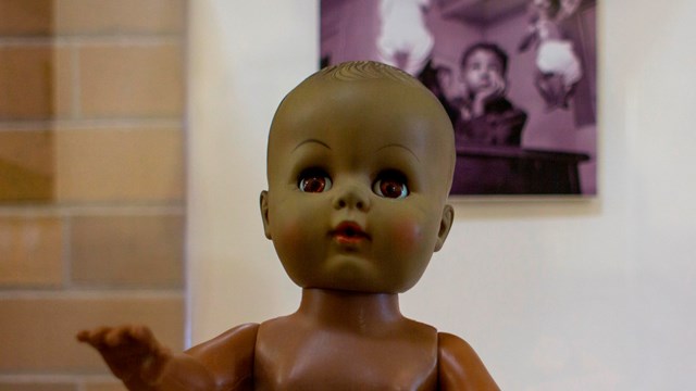 Brown doll in front of a photo of an African American child choosing between a while and brown doll.