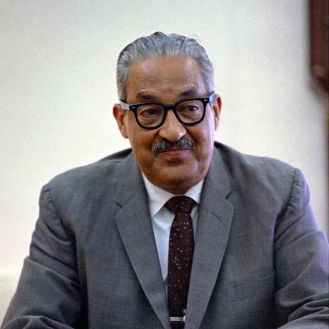 Thurgood Marshall sits in a grey suit leaning forward with his hands clasped.