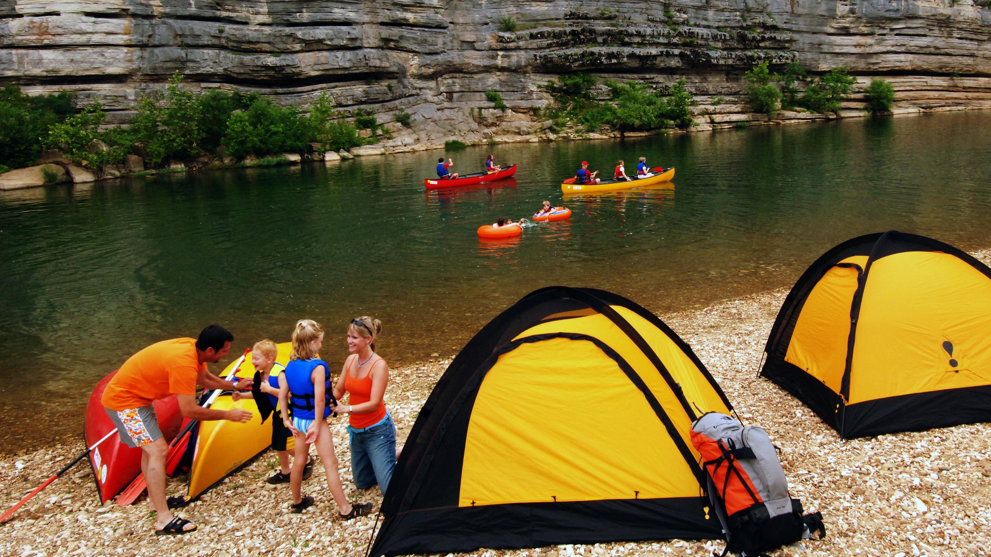 A family sets up tents on a gravel bar. Canoes float down the river past the gravel bar.