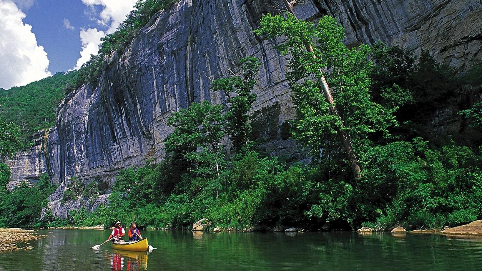 A kayak paddles in calm waters. A large bluff towers above.