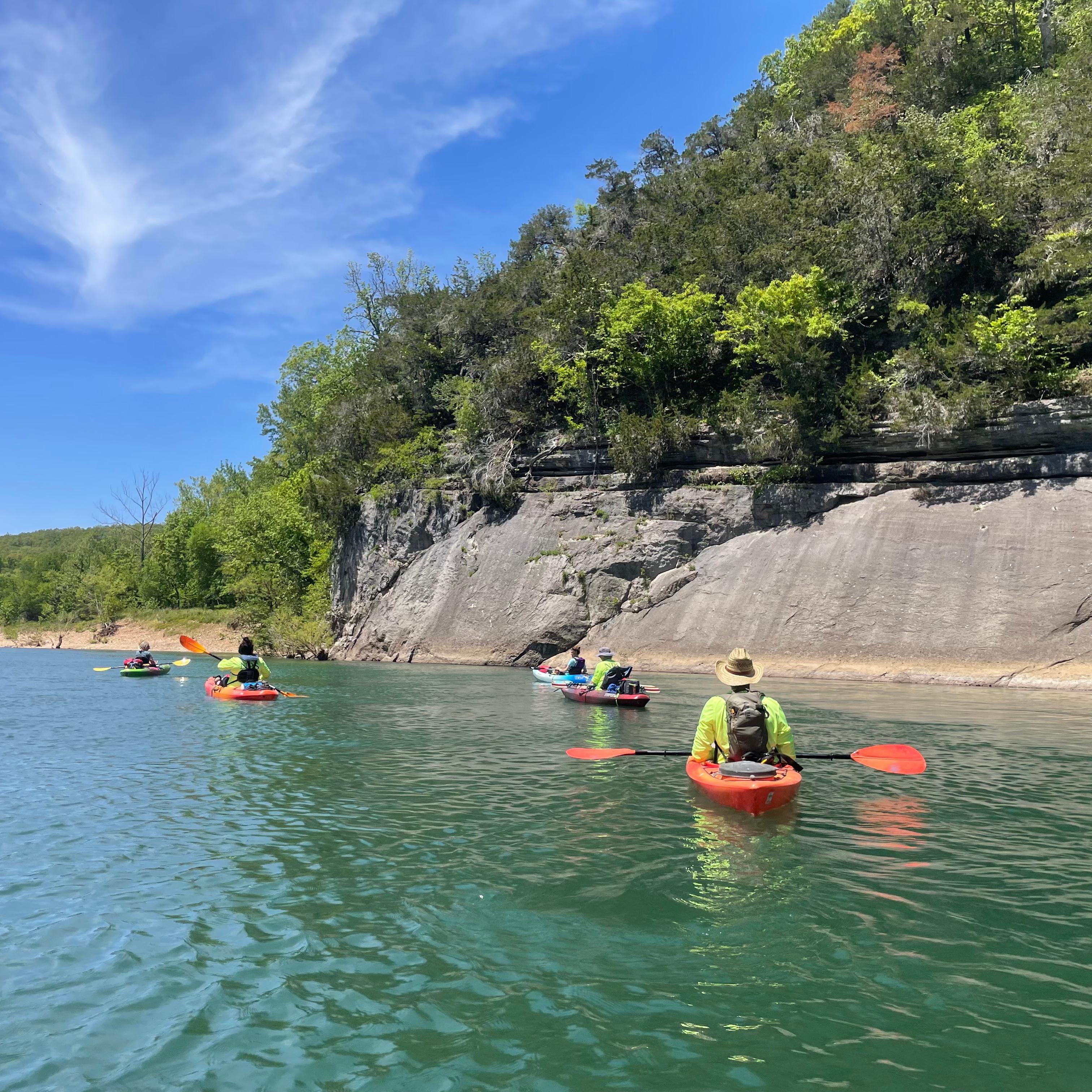 Kayakers floating on a blue river next to a rocky bluff.