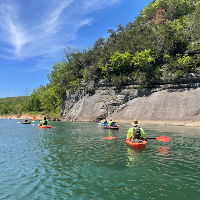 Kayakers floating on a blue river next to a rocky bluff.