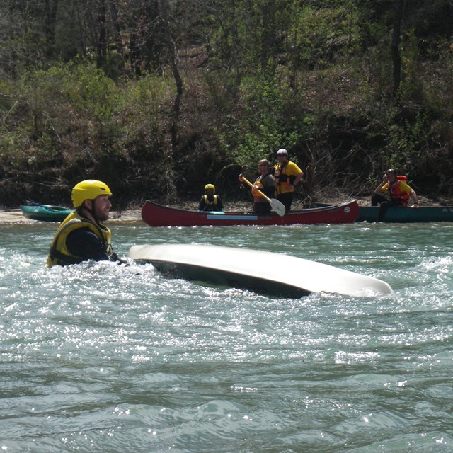 A man in a yellow helmet is in the river current with an upside down kayak. 