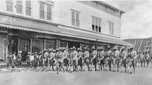 uniformed soldiers march in formation along a street with guns over their shoulders