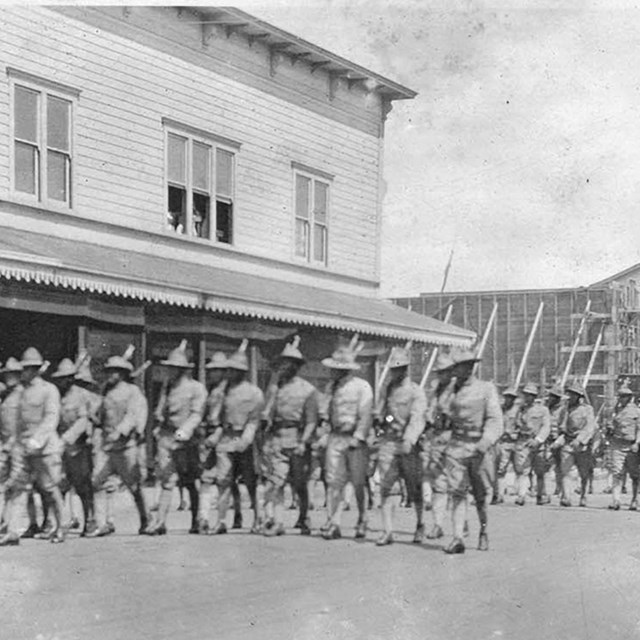 uniformed soldiers march in formation along a street with guns over their shoulders