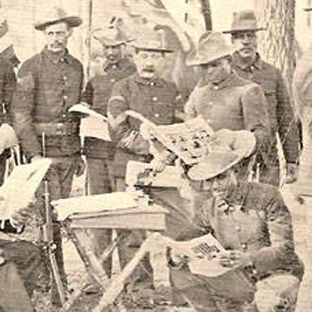 Several soldiers standing and sitting around a table and reading papers