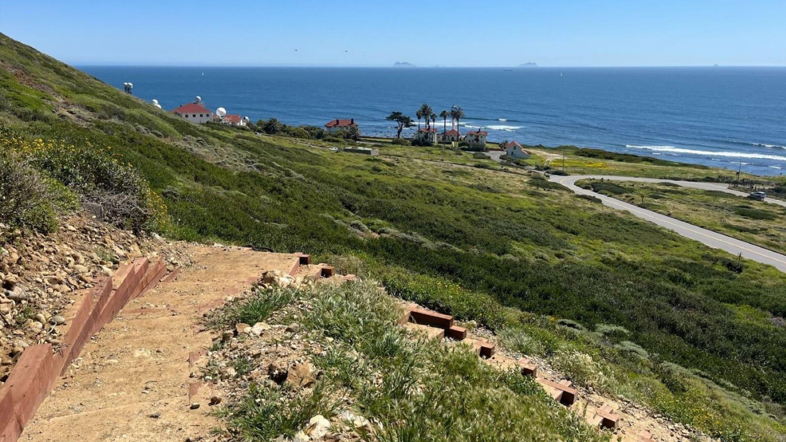 A  dirt path with switchbacks on a hillside with the ocean in the background.