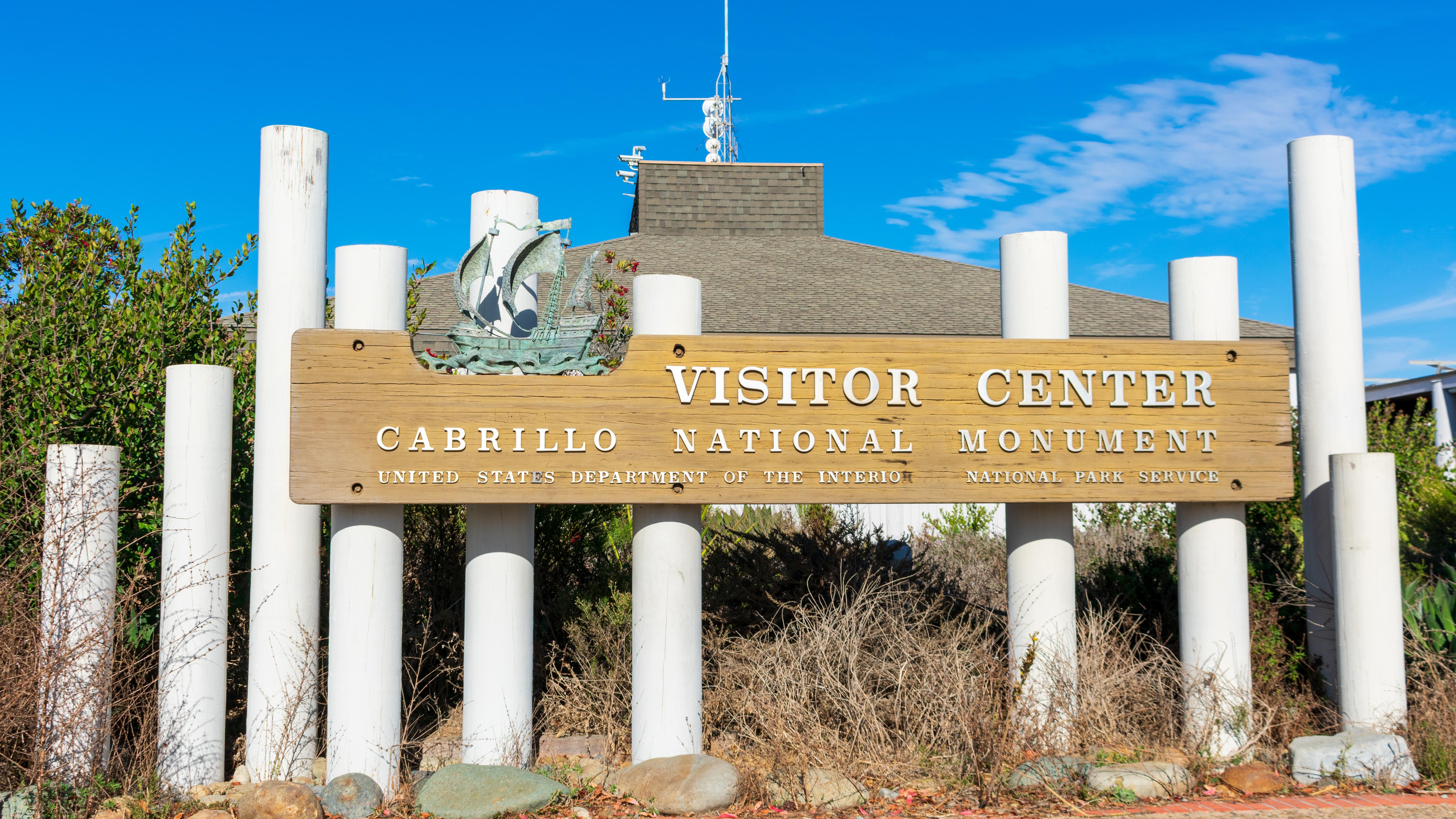 A wooden entrance sign with multiple posts and shrubs. Name of the park. 