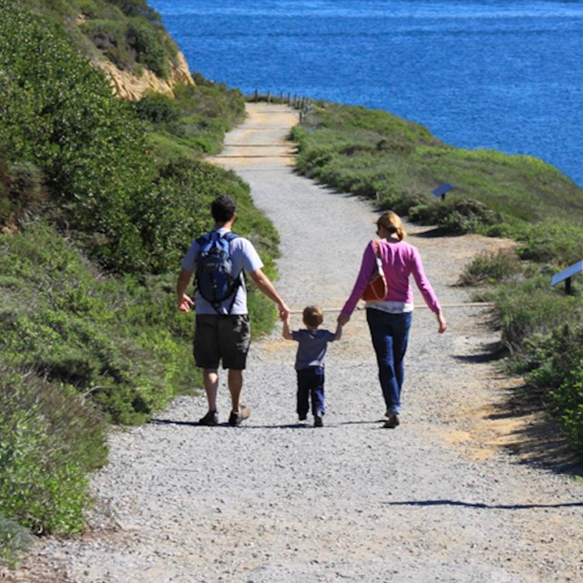 2 adults and a child walk hand-in-hand next to each other down a wide path west above the water.