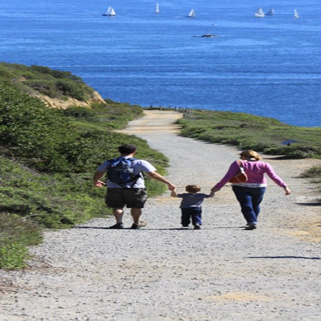 2 adults and a child walk hand-in-hand next to each other down a wide path west above the water.