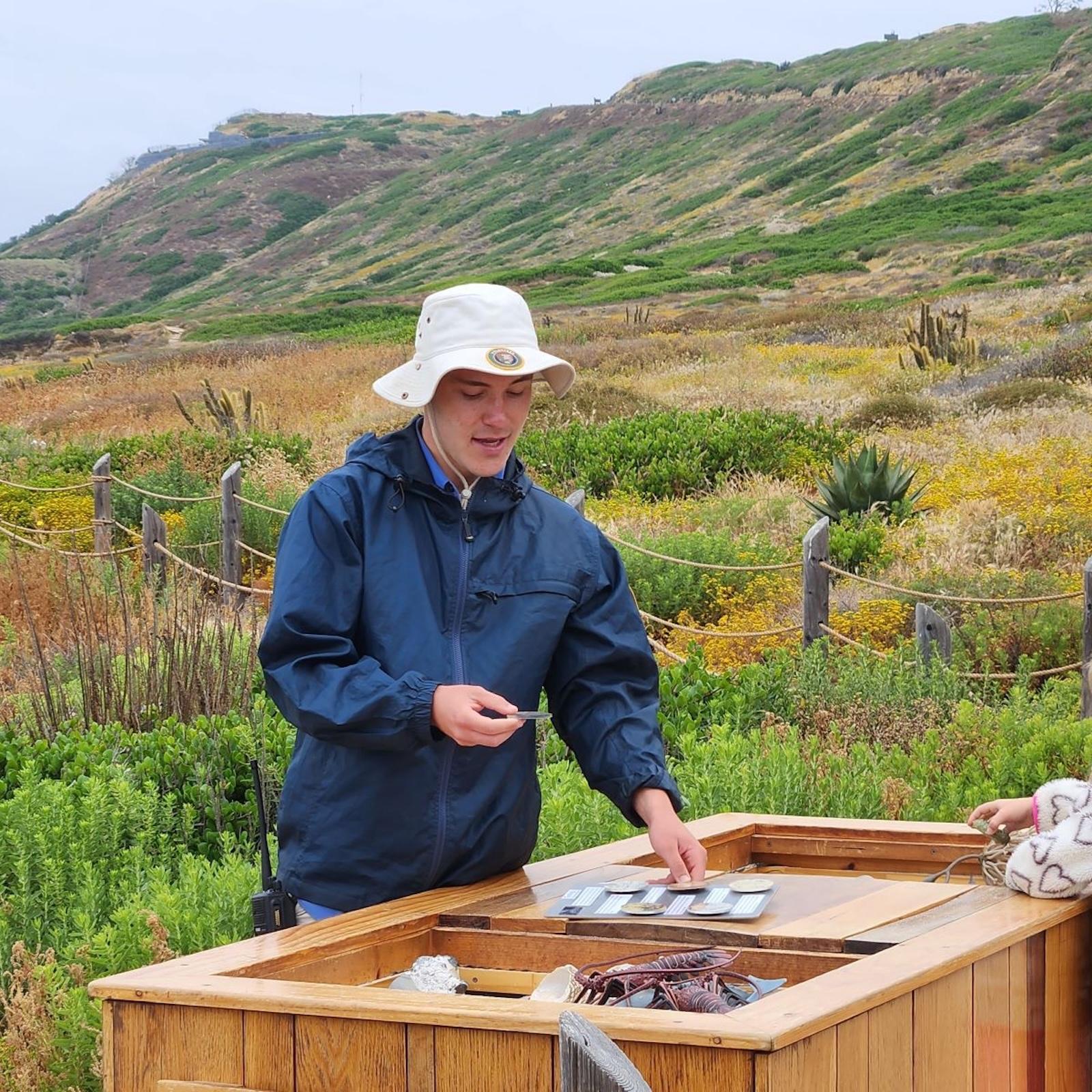 A person in a blue jacket and white sun hat stands at an outdoor wooden display table with cards