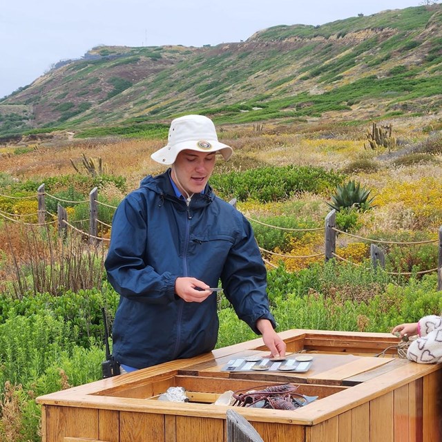 A person in a blue jacket and white sun hat stands at an outdoor wooden display table with cards
