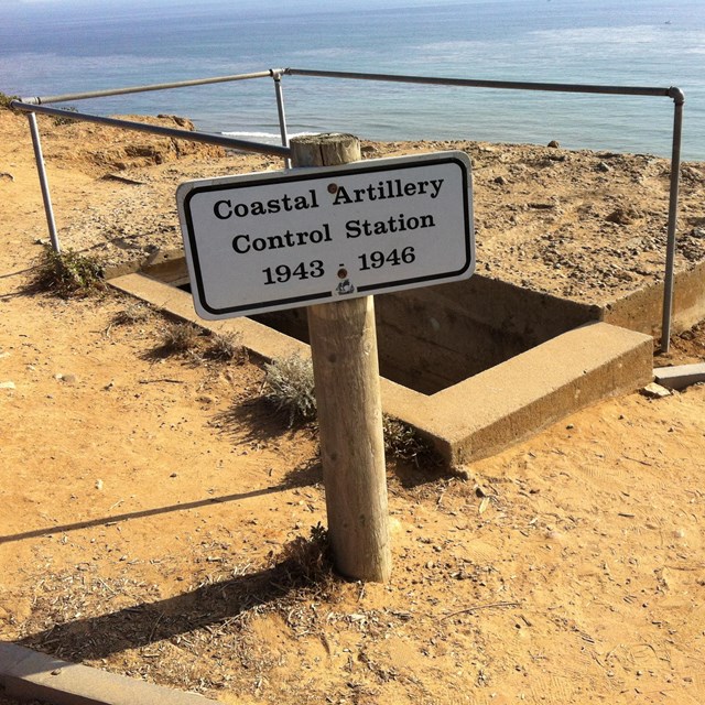 Concrete rectangle with shadow and metal rail surrounding in sandy ground. Ocean behind .