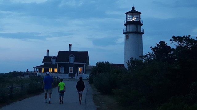 Three people walk up to a lighthouse with the tower light lit at dusk.