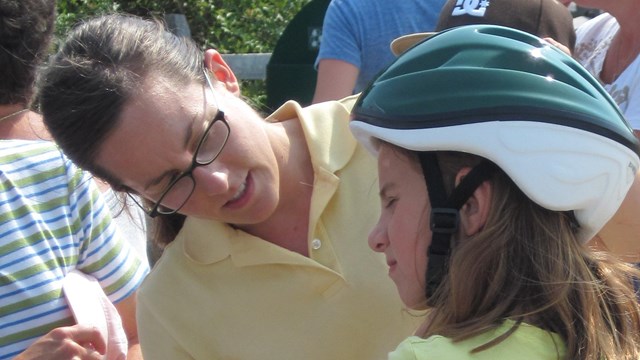 A young park visitor tries on a bike helmet while a volunteer helps her adjust it.