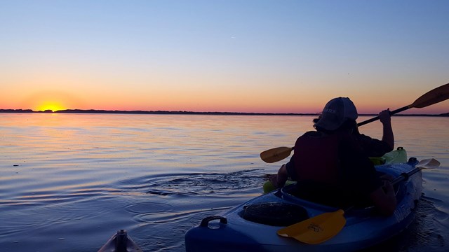 S person in a kayak on calm waters faces the setting sun.