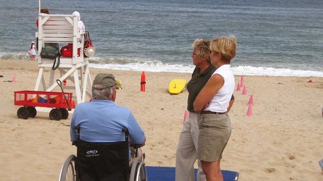 A man sits in a wheelchair at the end of a blue woven mat on a sandy beach talking to two women.