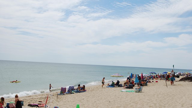 A crowd with colorful blankets and umbrellas enjoy beach day.