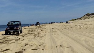 A black Jeep sits on a beach with people in swimsuits enjoying the sun.