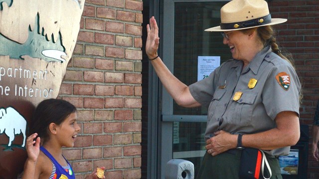 A ranger swears in two children as Junior Rangers outdoors in front of a large arrowhead logo.