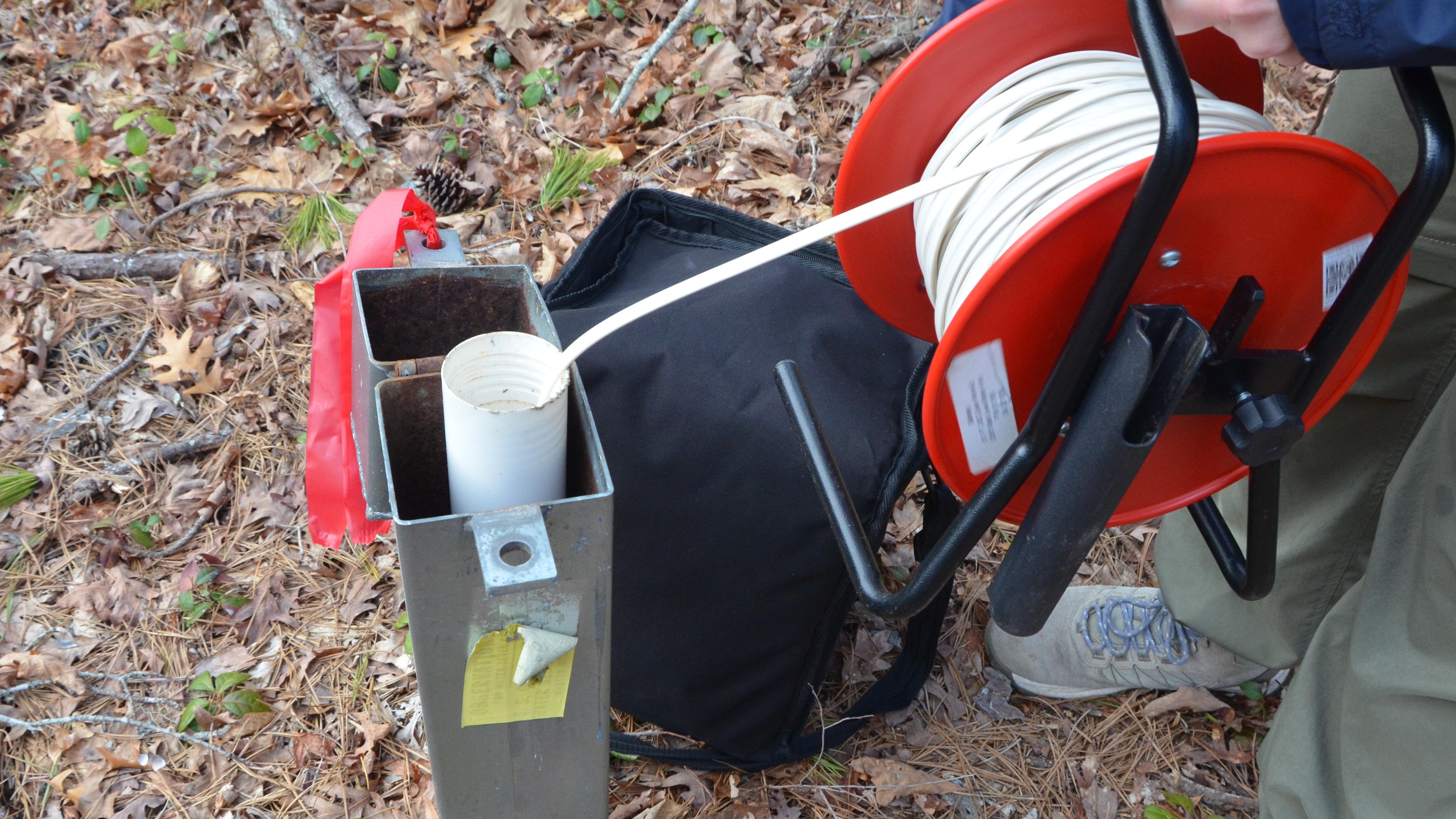 A technician&rsquo;s hands hold a red spool, with a line down the PVC pipe inside of a groundwater well, m