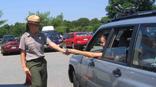 A ranger in uniform standing outside a small white wooden booth takes money from a person in a car.