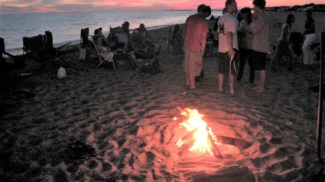 A group of people sit around a campfire on the beach.