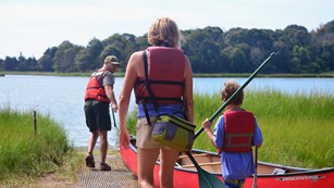 A ranger pulls a canoe down a ramp towards a pond, as a mother and son follow holding paddles.