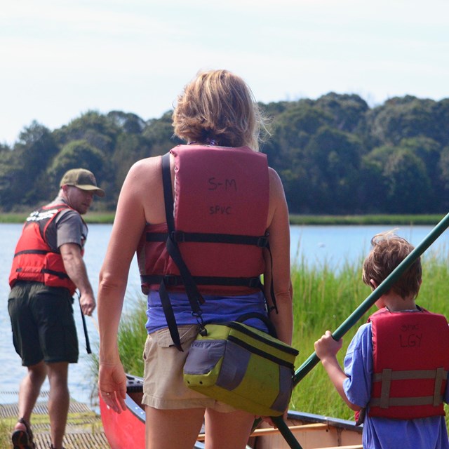 A ranger pulls a canoe down a ramp towards the water while a mother and son with paddles follow him.