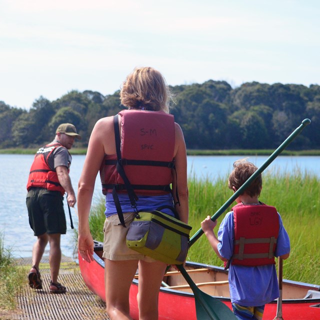 A ranger pulls a canoe down a ramp towards a pond, as a mother and son follow holding paddles.