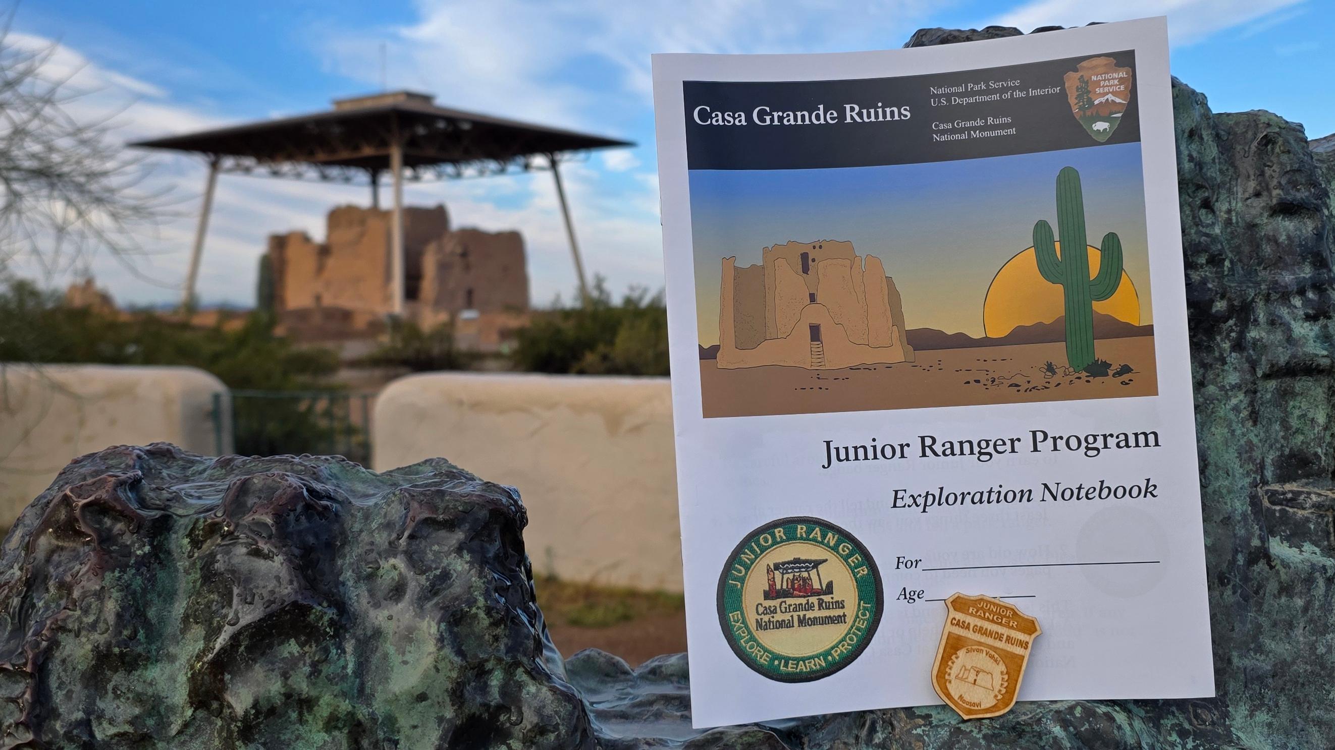 A Junior Ranger Badge and Book in creosote with a large adobe structure in the background. 