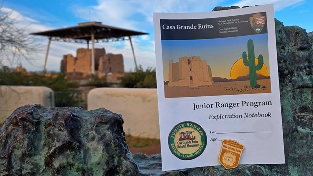 A Junior Ranger Badge and Book in creosote with a large adobe structure in the background. 