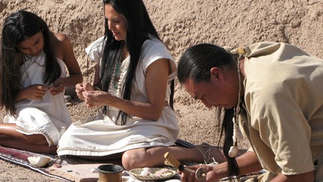 scene from park film with 3 costumed actors making jewelry with stone tools