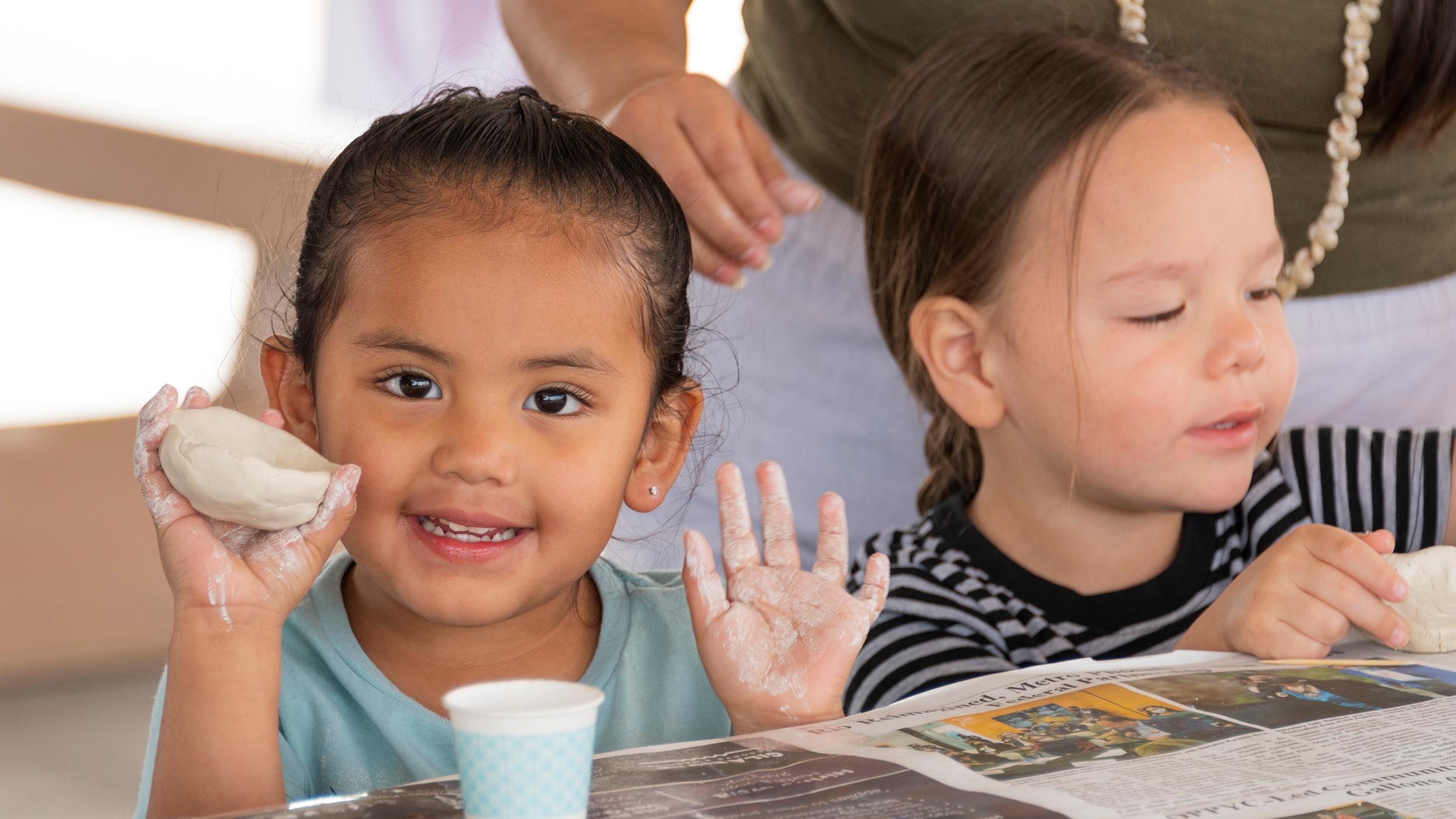 A student holds up a clay pinch pot while another student works on a piece of clay 