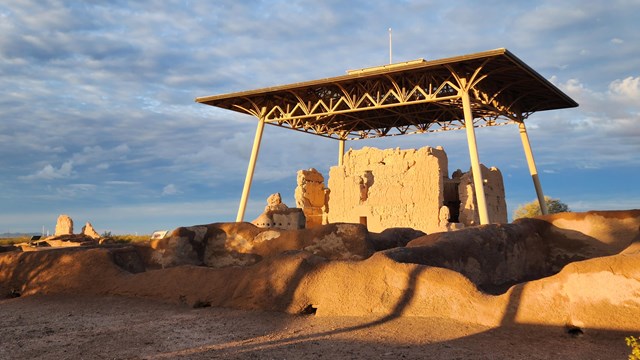 A large ancient adobe structure with a protective metal roof structure lit by the sun at sunrise